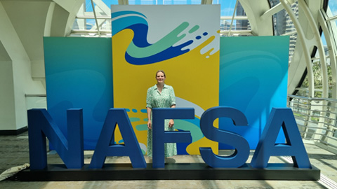 Person standing behind large blue 'NAFSA' letters, with colorful abstract panels and natural light in the background.